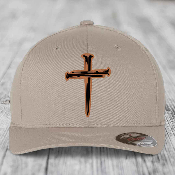 Beige baseball cap with a cross of nails patch on front, displayed on wooden surface.