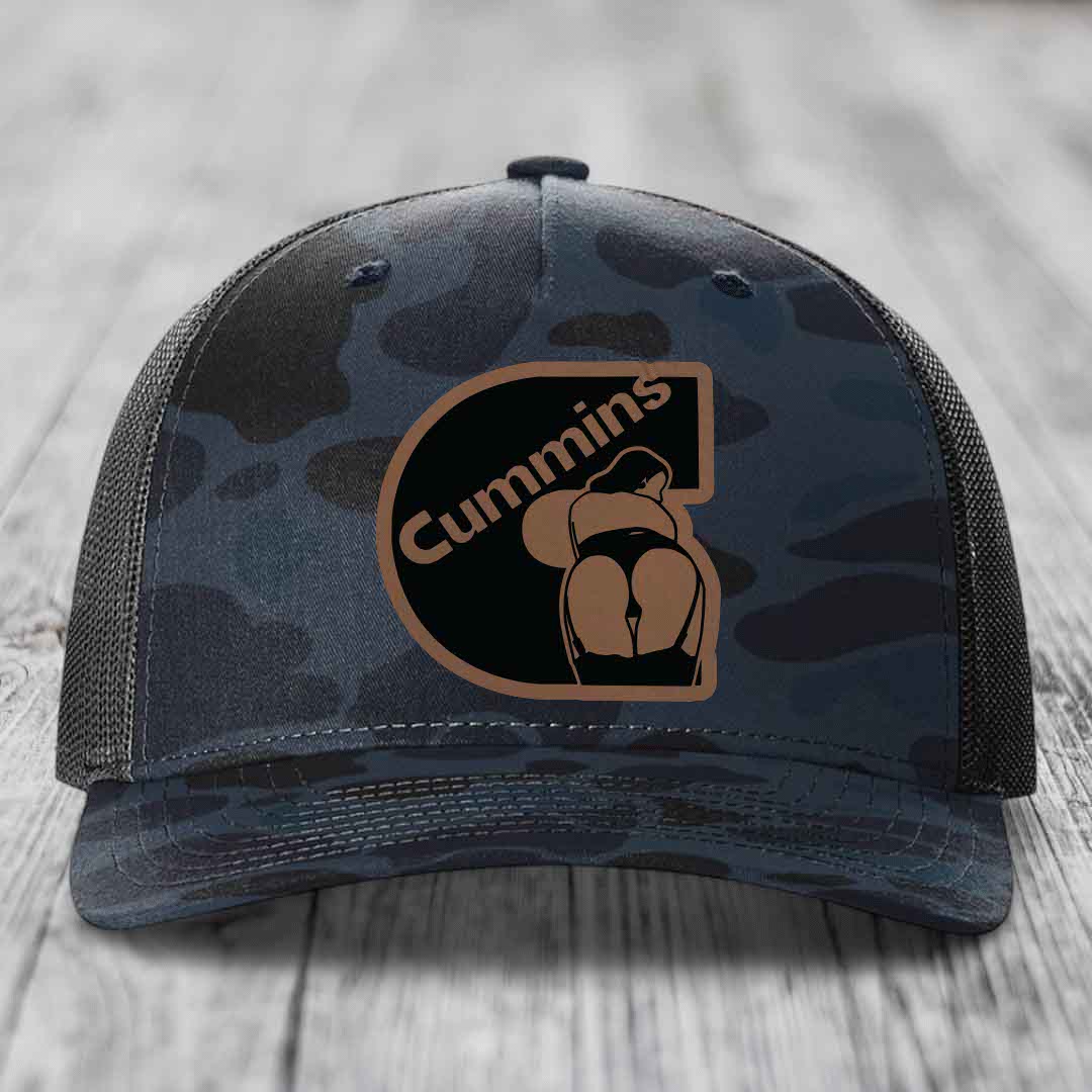 Camouflage trucker hat with Cummins logo patch on front, placed on wooden surface.
