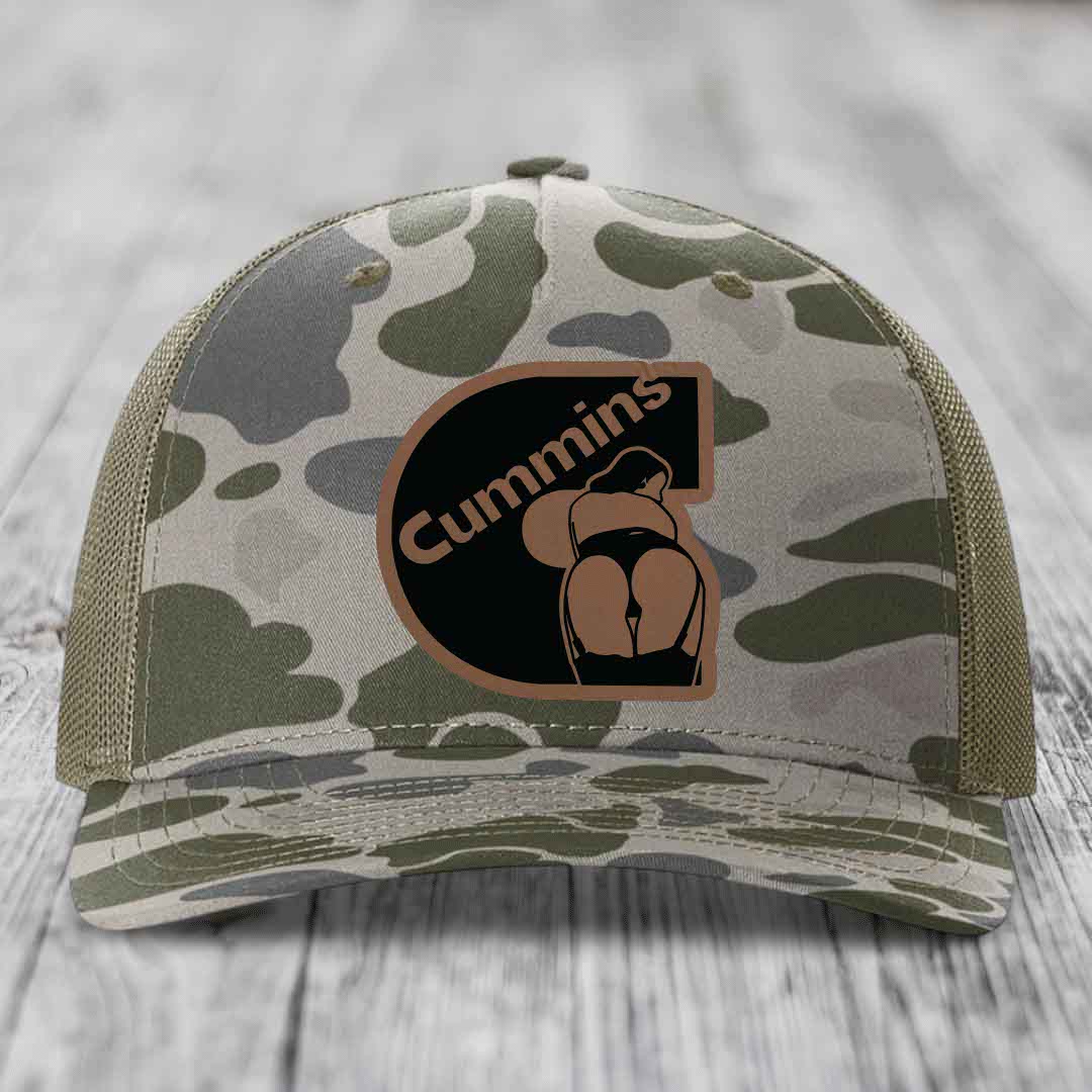 Camouflage trucker hat with Cummins logo patch on front, placed on wooden surface.
