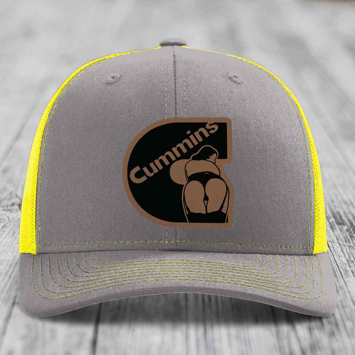 Brown trucker hat with black and tan Cummins logo patch on front, white mesh back.