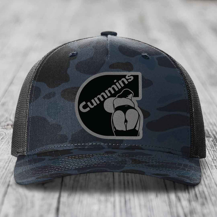Camouflage trucker hat with black Cummins logo patch on front, sitting on wooden surface.