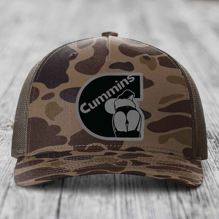 Camouflage trucker hat with black Cummins logo patch on front.