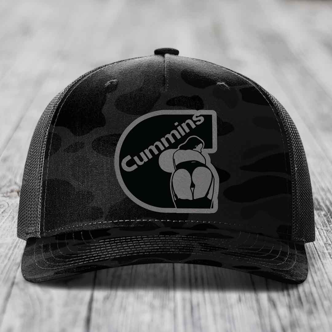 Camouflage trucker hat with black Cummins logo patch on front, gray mesh back, sitting on wooden surface.