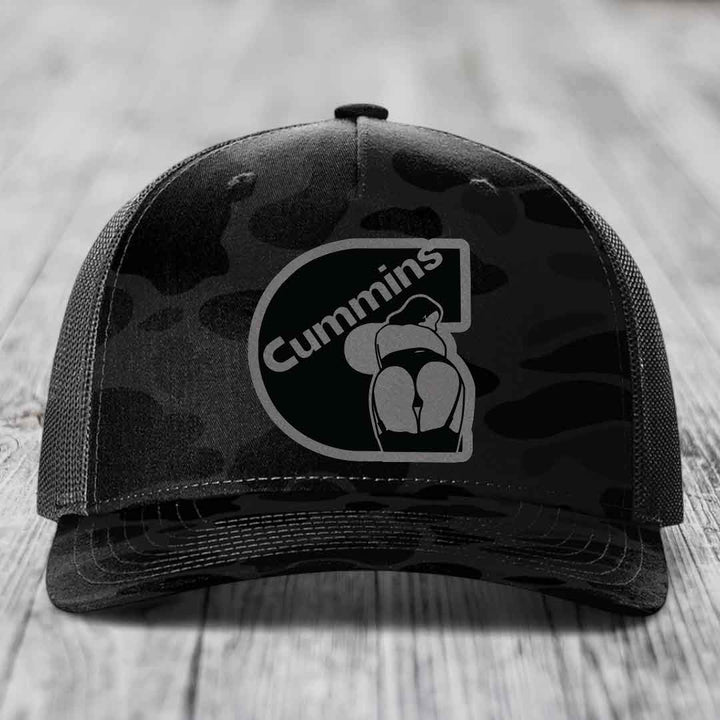 Camouflage trucker hat with black Cummins logo patch on front, gray mesh back, sitting on wooden surface.