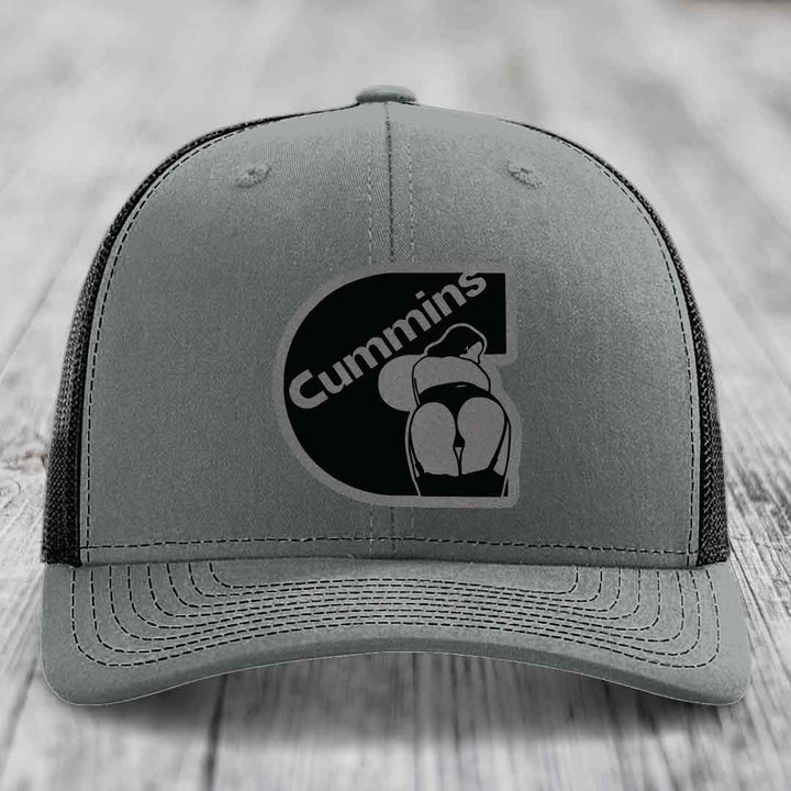Grey trucker hat with black Cummins logo patch on front, white mesh back, and white stitching.
