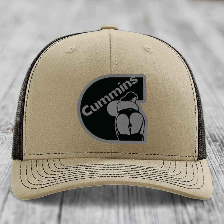 Tan trucker hat with black Cummins logo patch on front, white mesh back, and white stitching.