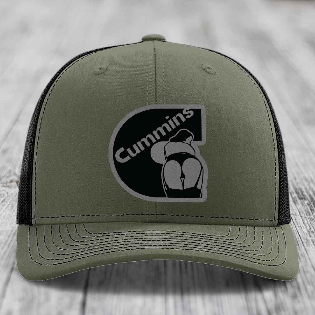 Black trucker hat with grey Cummins logo patch on front, resting on wooden surface.