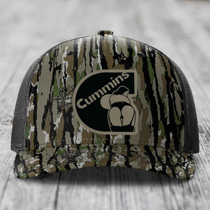 Black camouflage trucker hat with Cummins logo patch on front, resting on wooden surface.