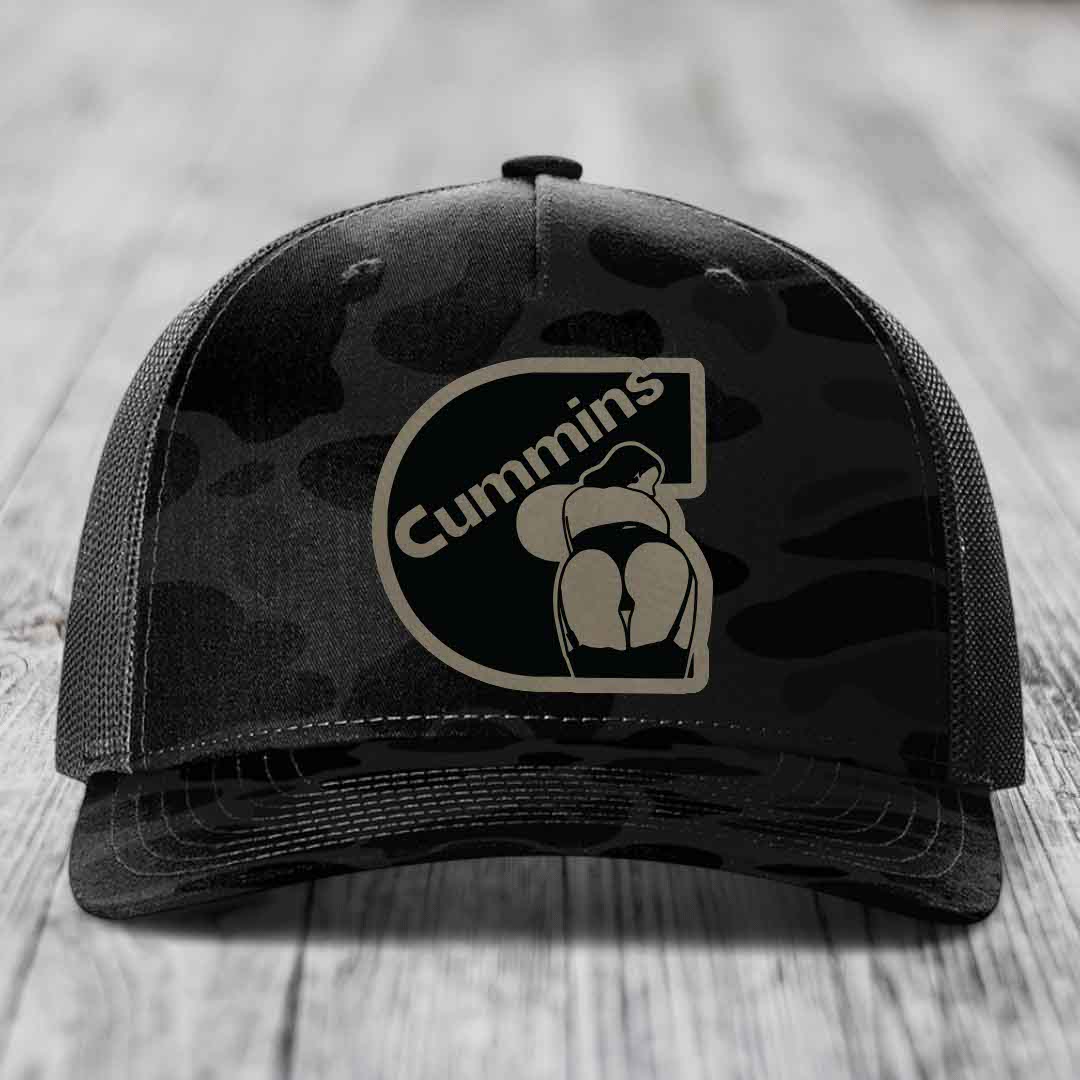 Camouflage trucker hat with black Cummins logo patch on front, gray mesh back, sitting on wooden surface.