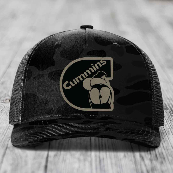 Camouflage trucker hat with black Cummins logo patch on front, gray mesh back, sitting on wooden surface.