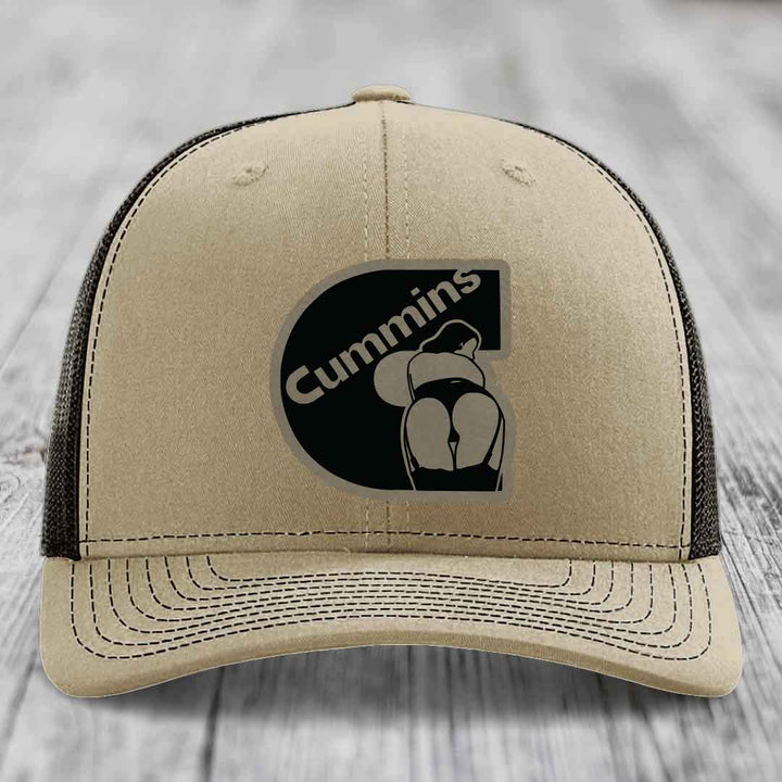 Tan trucker hat with black Cummins logo patch on front, white mesh back and stitching.