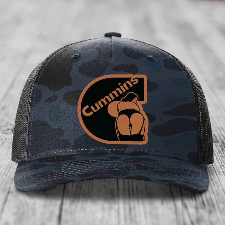 Camouflage trucker hat with Cummins logo patch on front, placed on wooden surface.