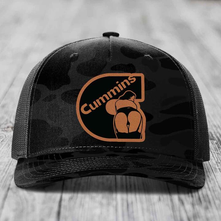 Camouflage trucker hat with Cummins logo patch on front, placed on wooden surface.