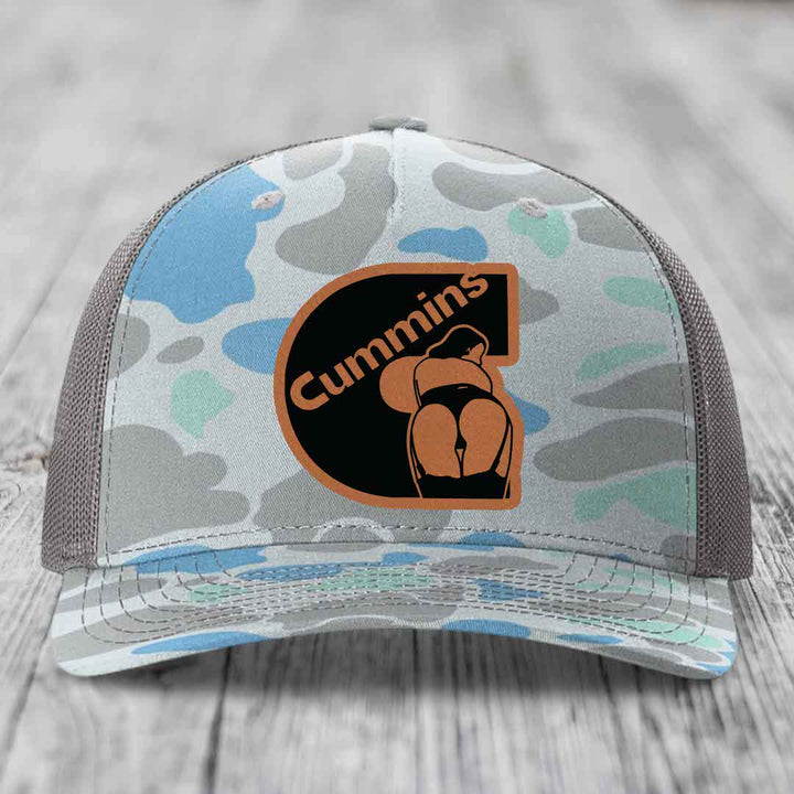 Camouflage trucker hat with Cummins logo patch on front, black mesh back, sitting on wooden surface.