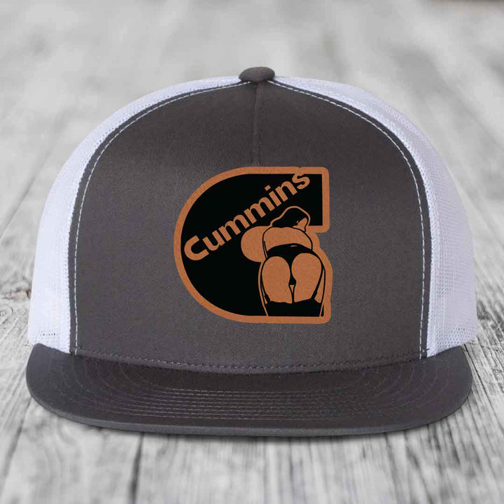 Black snapback trucker hat with grey Cummins logo patch on front, resting on wooden surface.