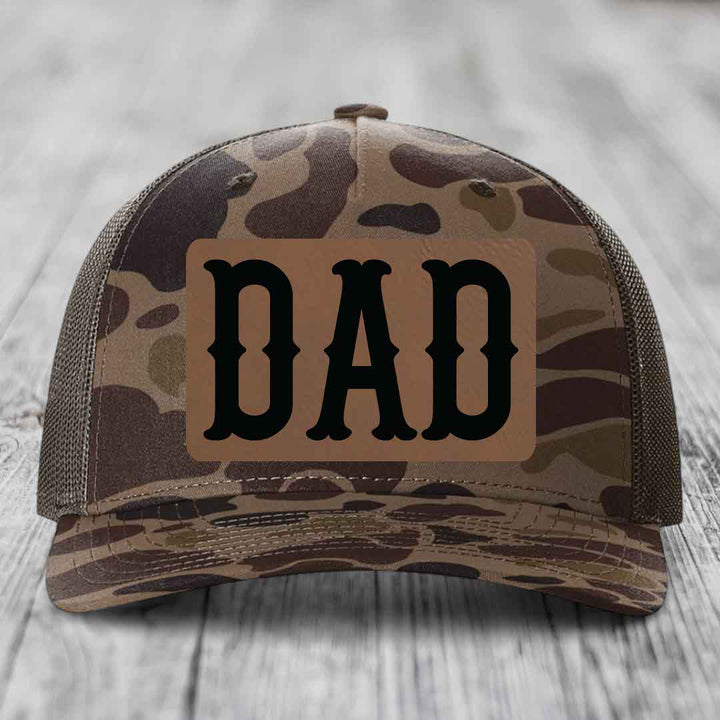 Camouflage trucker hat with brown leather patch that says DAD in black letters.