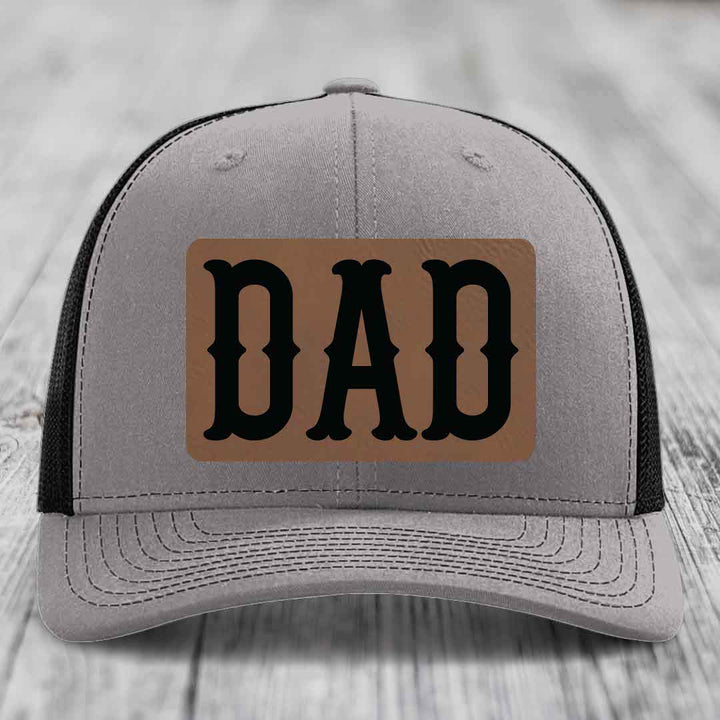 Grey trucker hat with blue mesh sides and a brown leather patch that says DAD in black letters.