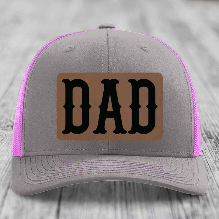 Grey trucker hat with yellow mesh sides and stitching, featuring a brown leatherette patch with black &