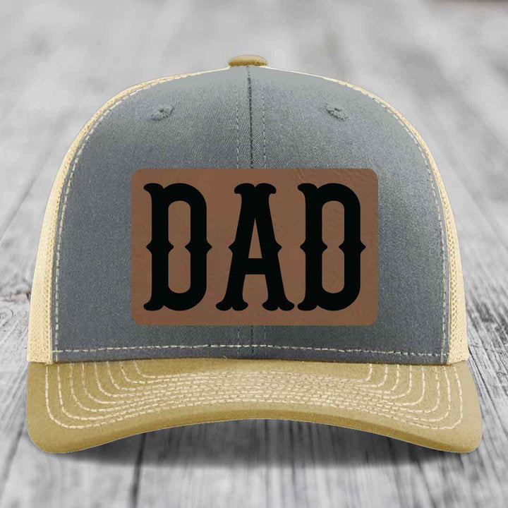 Grey trucker hat with black mesh back and brown leather patch that says DAD in black letters.
