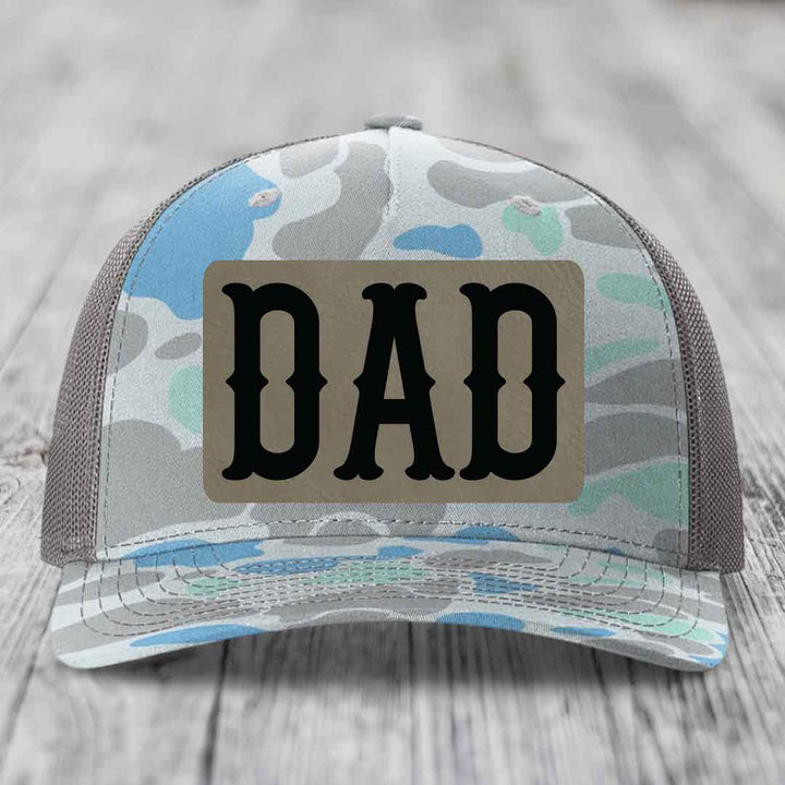 Camouflage trucker hat with a brown leatherette patch that says &