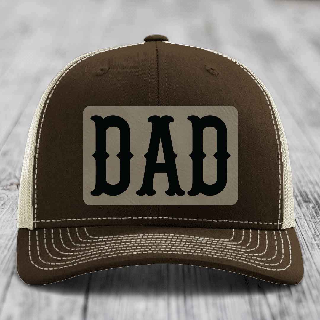 Tan trucker hat with black mesh back and a grey leatherette patch that says DAD in black letters.