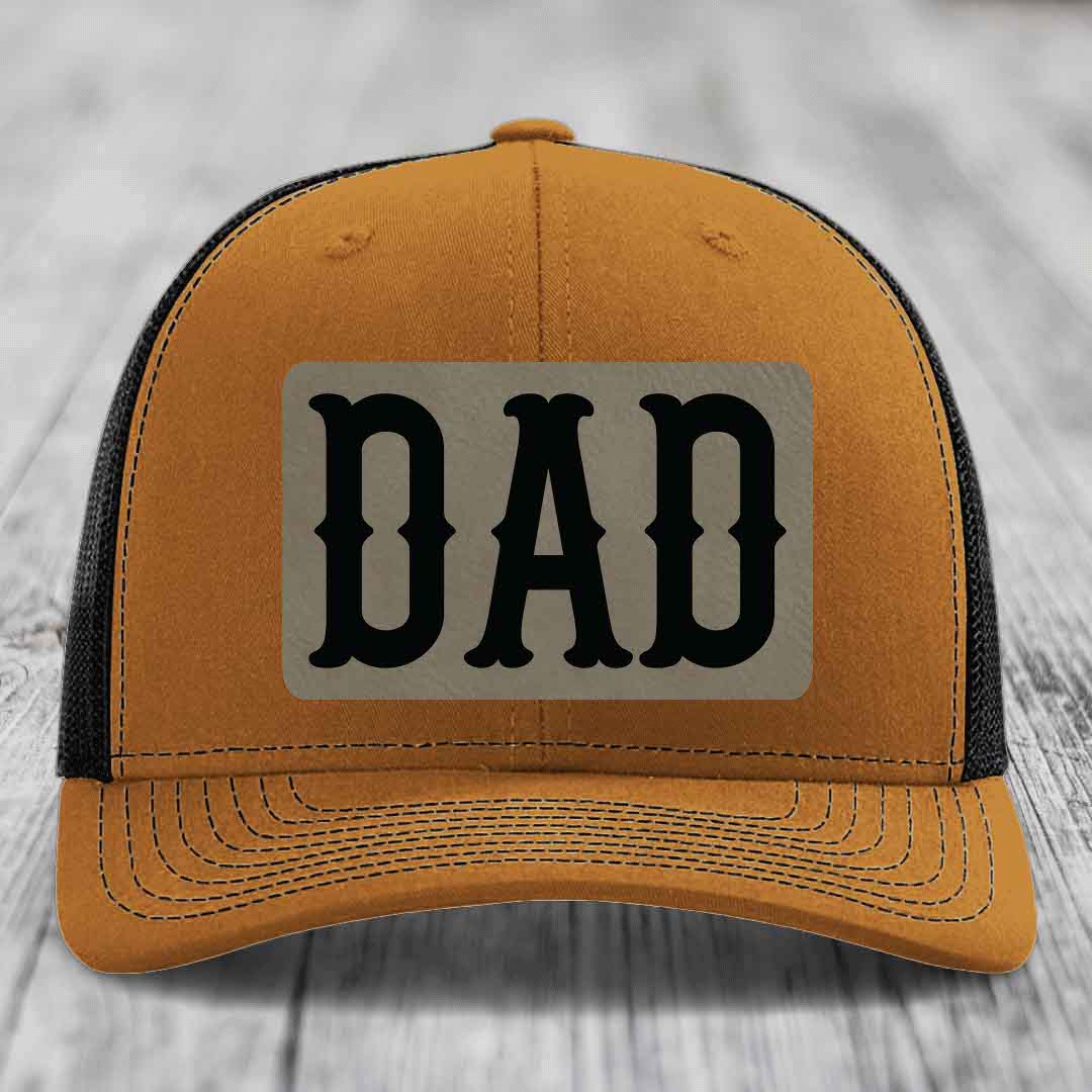 Grey trucker hat with black mesh back and a beige leatherette patch that says DAD in black letters.