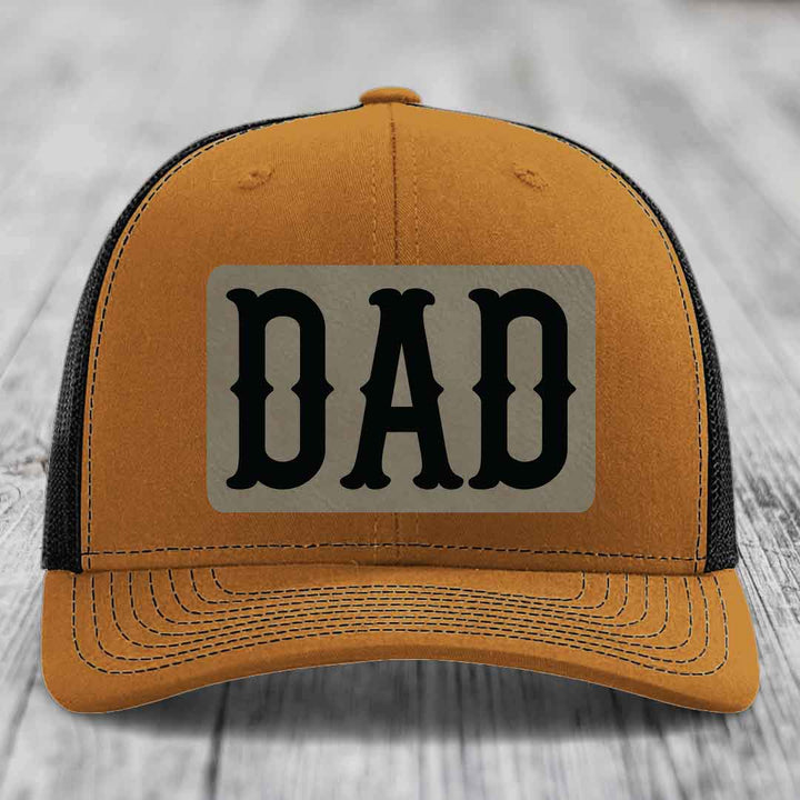 Grey trucker hat with black mesh back and a beige leatherette patch that says DAD in black letters.