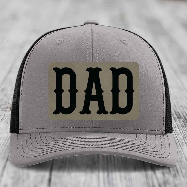 Grey trucker hat with blue mesh sides and a beige leatherette patch that says DAD in black letters.