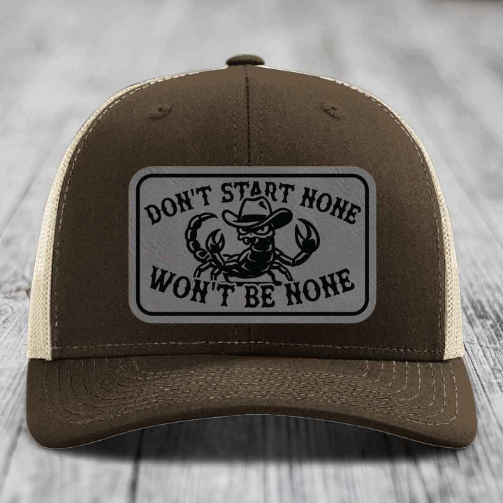 Brown trucker hat with beige mesh back and a rectangular leatherette patch featuring a scorpion in a cowboy hat and the text &
