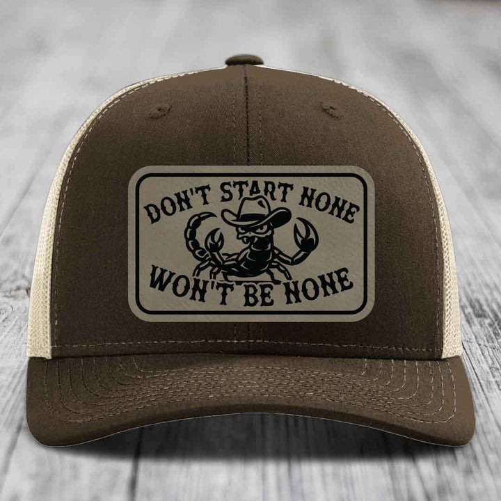 Brown trucker hat with beige mesh back and a rectangular leather patch featuring a scorpion wearing a cowboy hat and the text &