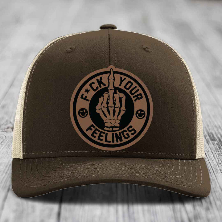 Brown trucker hat with beige mesh back and a circular leather patch showing skeleton hand making rude gesture with text &