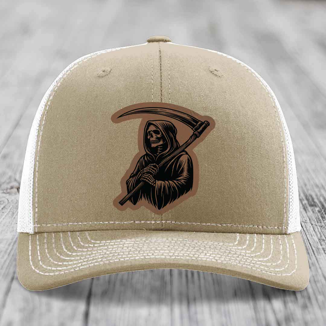 Tan trucker hat with black Grim Reaper patch holding scythe, white mesh back and stitching.