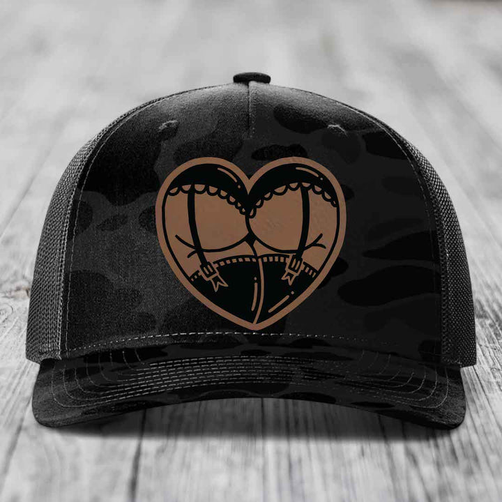 Black camouflage trucker hat with heart-shaped garter belt graphic in tan and black.