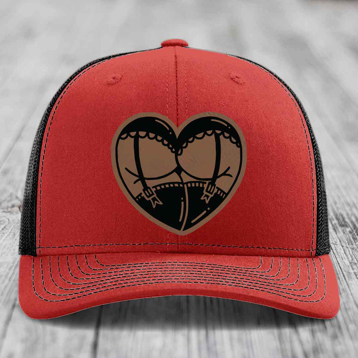 Red trucker hat with black mesh back and a heart-shaped leather patch featuring a garter belt design.