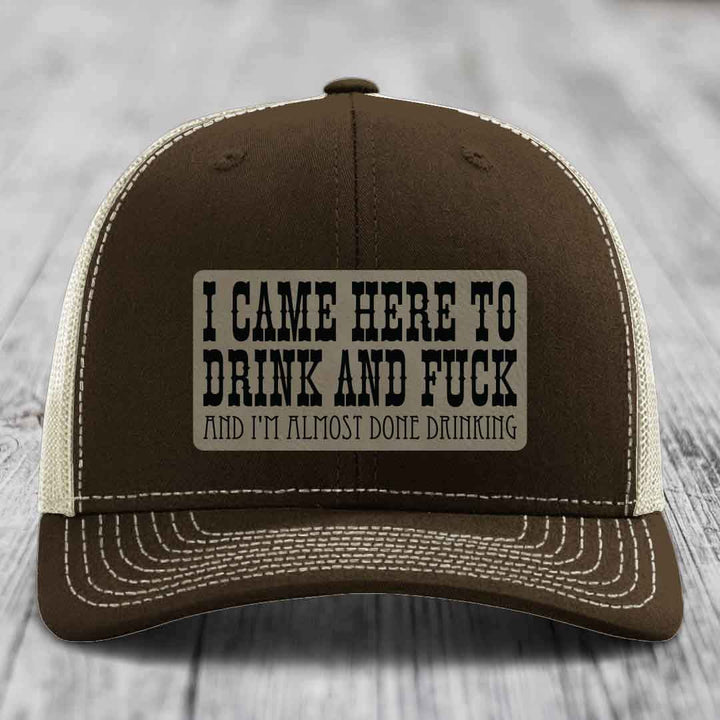 Brown trucker hat with white mesh back and stitched details, featuring a rectangular leatherette patch with bold black text reading &