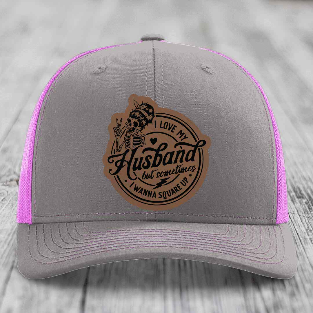 Grey trucker hat with pink mesh, featuring a brown leatherette patch of a skeleton making peace sign with text &
