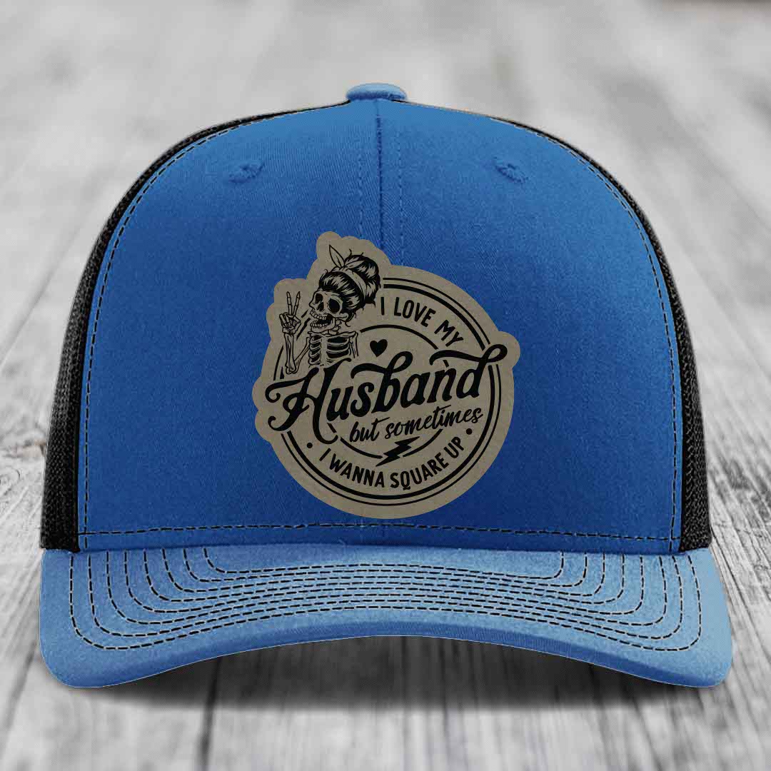 Blue trucker hat with black mesh back and a beige leatherette patch featuring a skeleton graphic and text.