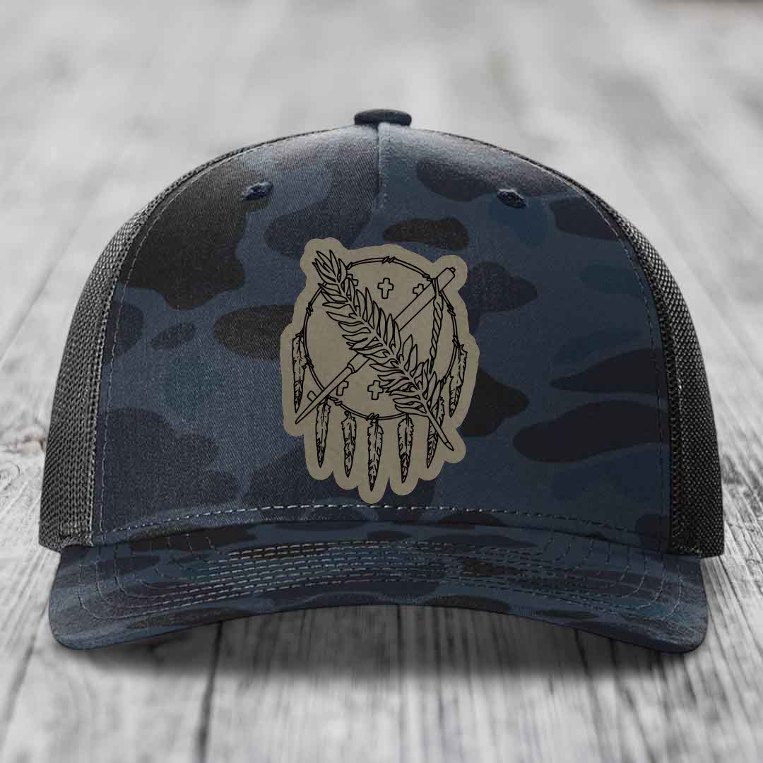 Camouflage trucker hat with a beige leatherette patch featuring a dreamcatcher design with feathers and crosses.