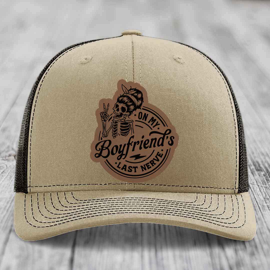 Tan trucker hat with black mesh back and a brown leather patch featuring a skeleton making a peace sign and text &
