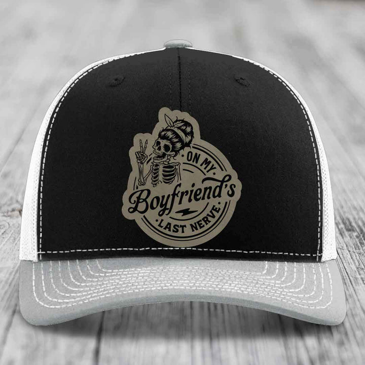 Black and grey trucker hat with a beige leatherette patch featuring a skeleton girl making a peace sign and text &