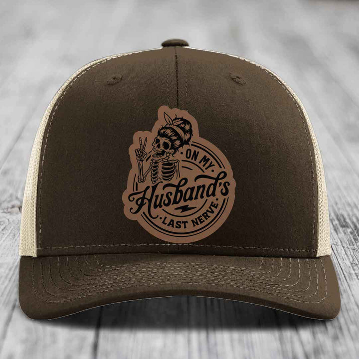 Brown trucker hat with beige mesh back and a tan leather patch featuring a skeleton woman making a peace sign with the text &