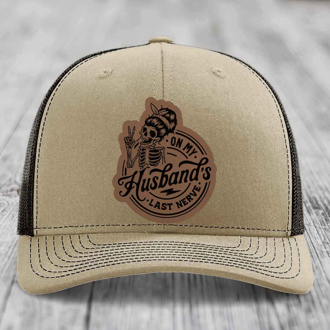 Beige trucker hat with black mesh back and a brown leather patch featuring a skeleton woman making a peace sign and the text &