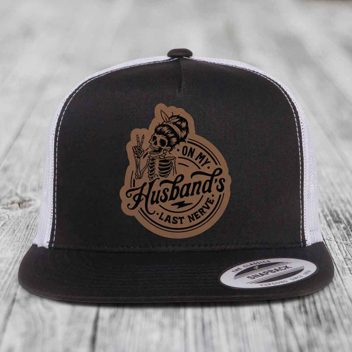 Black and white trucker hat with a brown leatherette patch featuring a skeleton woman making a peace sign and the text &