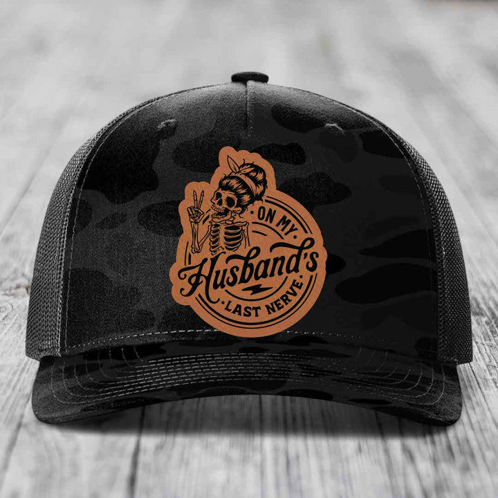 Black camouflage trucker hat with a tan leatherette patch featuring a skeleton woman making a peace sign and the text &