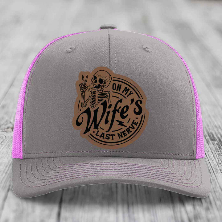 Grey trucker hat with pink mesh, featuring a brown leatherette patch of a skeleton making peace sign with text &
