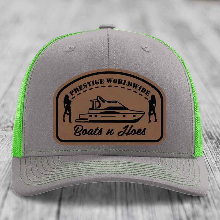 Grey trucker hat with neon green mesh and a brown leather patch featuring a boat and two silhouettes.