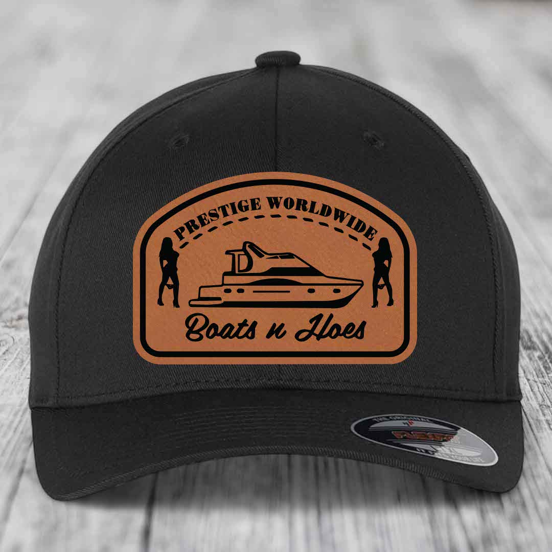 Black baseball cap with brown leather patch featuring yacht and two silhouettes, text reads &