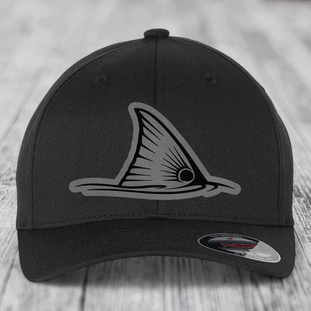 Black baseball cap with grey shark fin patch on front, sitting on wooden surface.