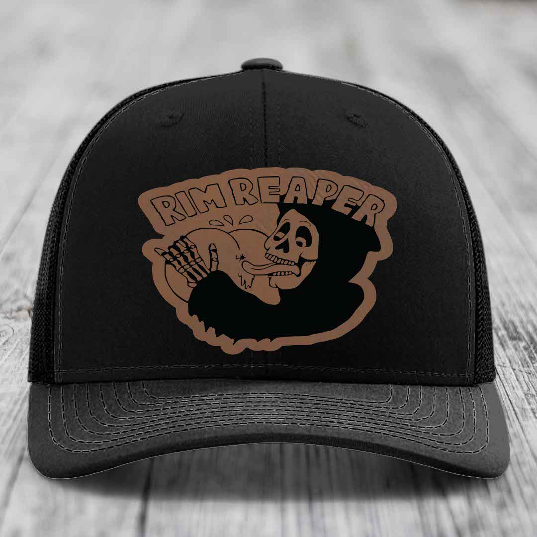 Black trucker hat with brown leatherette patch featuring skull holding baseball, text reads &