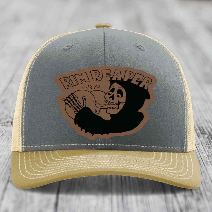 Grey front panel trucker hat with tan mesh back and brim, featuring a brown leather patch with a skull holding a baseball and &